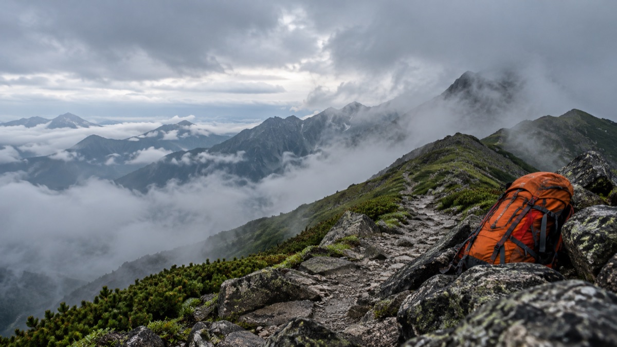 雲のかかる北海道の山並みと登山道に置かれたオレンジ色のザックのイメージ